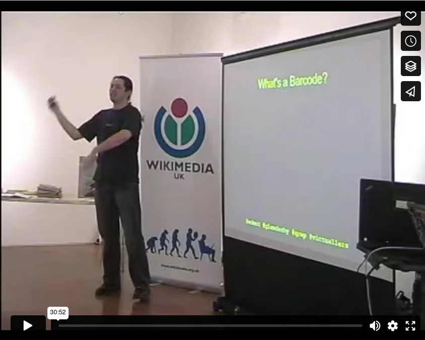 Capture from a video. Terence Eden stands in front of a projection screen. There is a Wikimedia banner behind him.