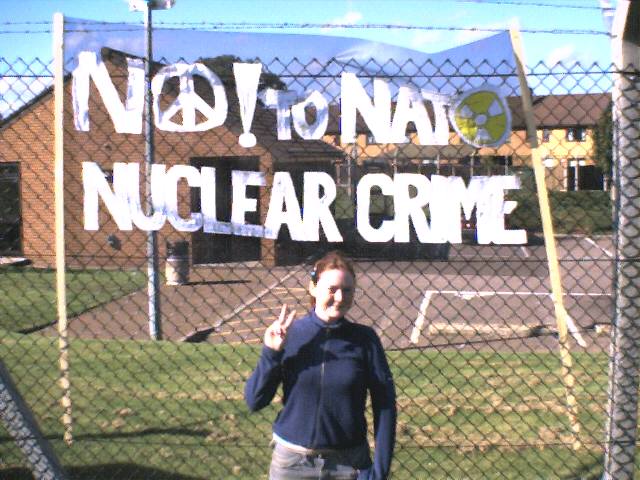 A young woman flashes the peace sign in front of a metal fence. There is a sign saying No to NATA Nuclear Crime.