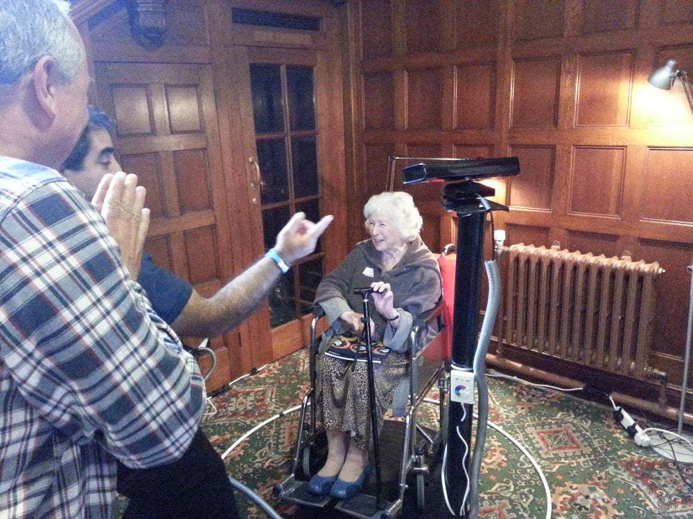 An old woman in a wheelchair smiles as she is 3D scanned.