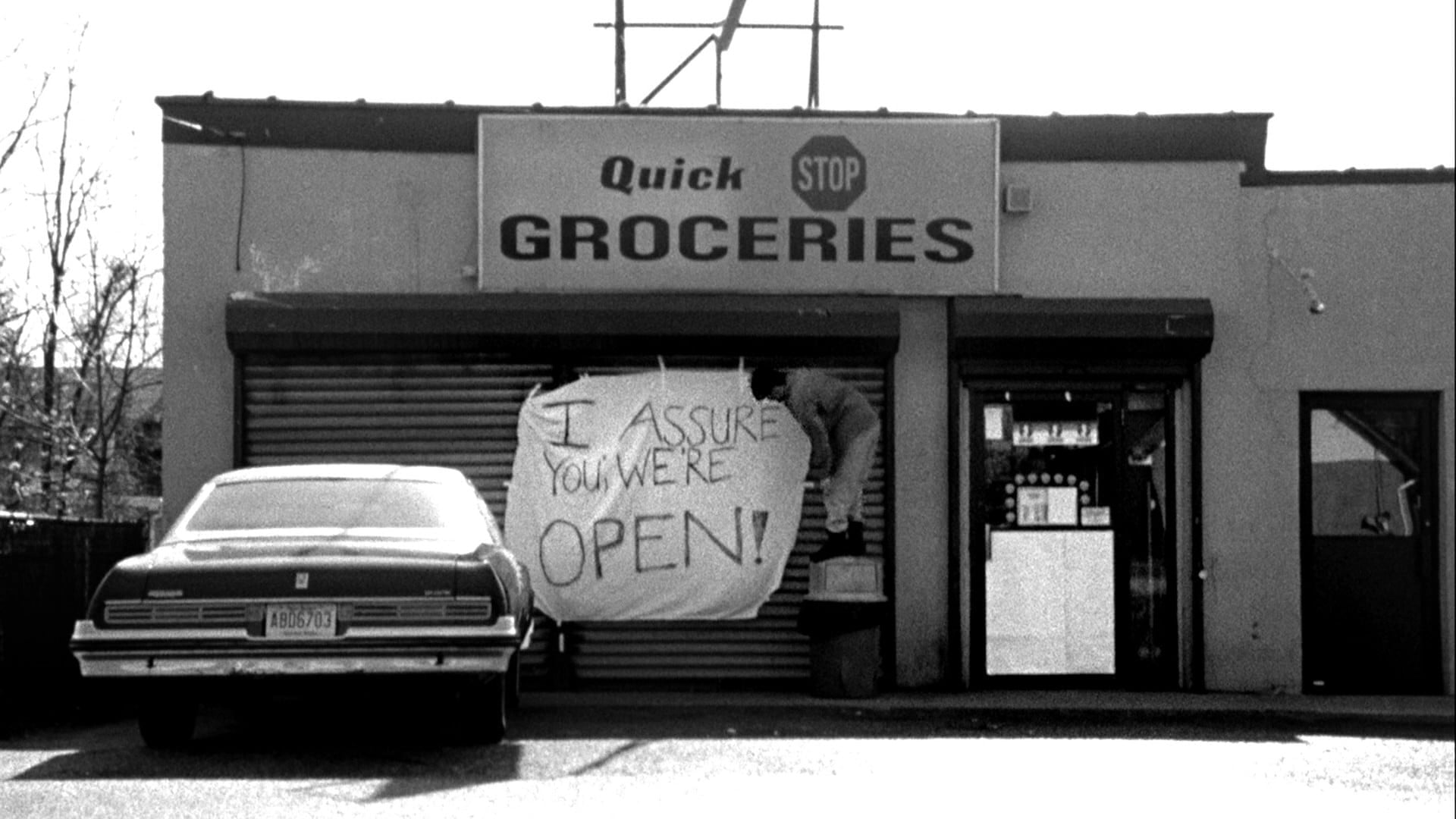 Still from the movie "Clerks". A convenience store has the shutters closed. A handwritten sign says "I assure you we're open!"