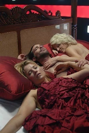 Two lady robots lay entwined with a bloke in red sheets.