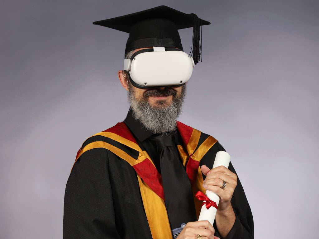Photo of a bearded man, wearing full academic dress and a VR headset.