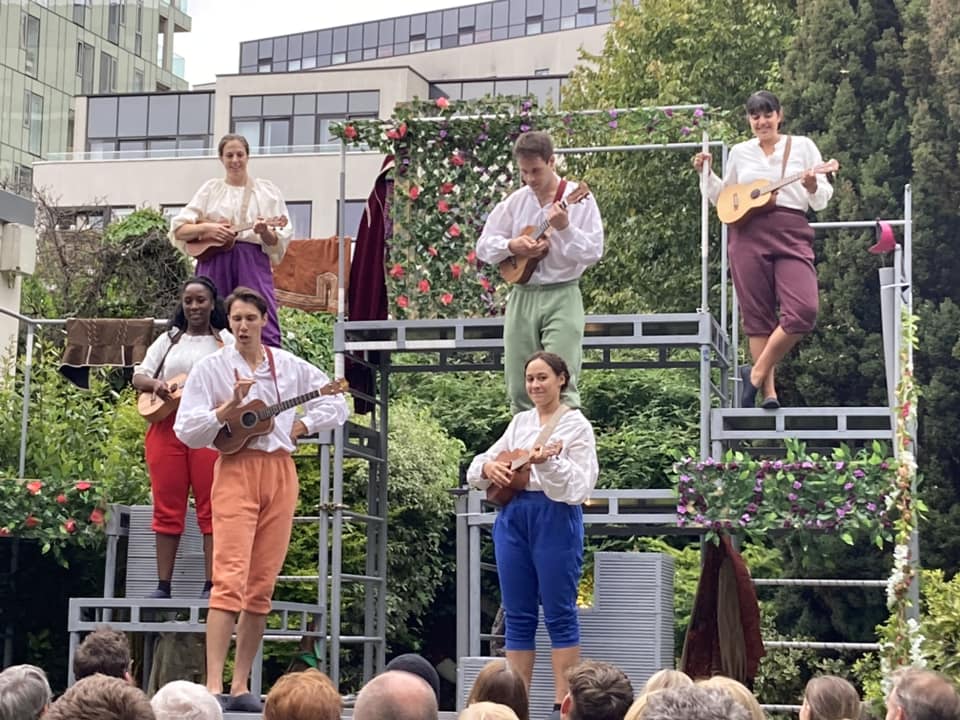 Six actors stand on a stage made out of scaffolding. They are all playing ukuleles.