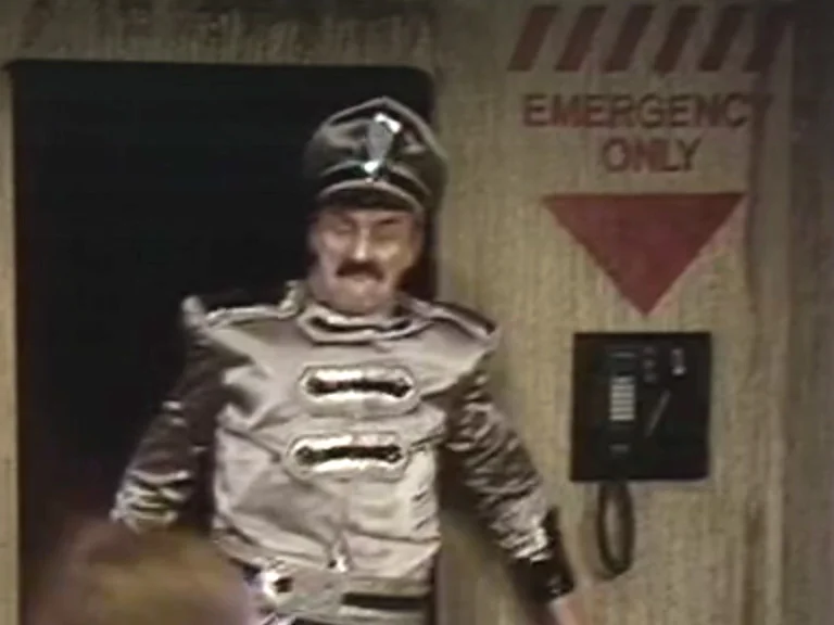 A man in a silver uniform stands next to a wall-mounted phone.