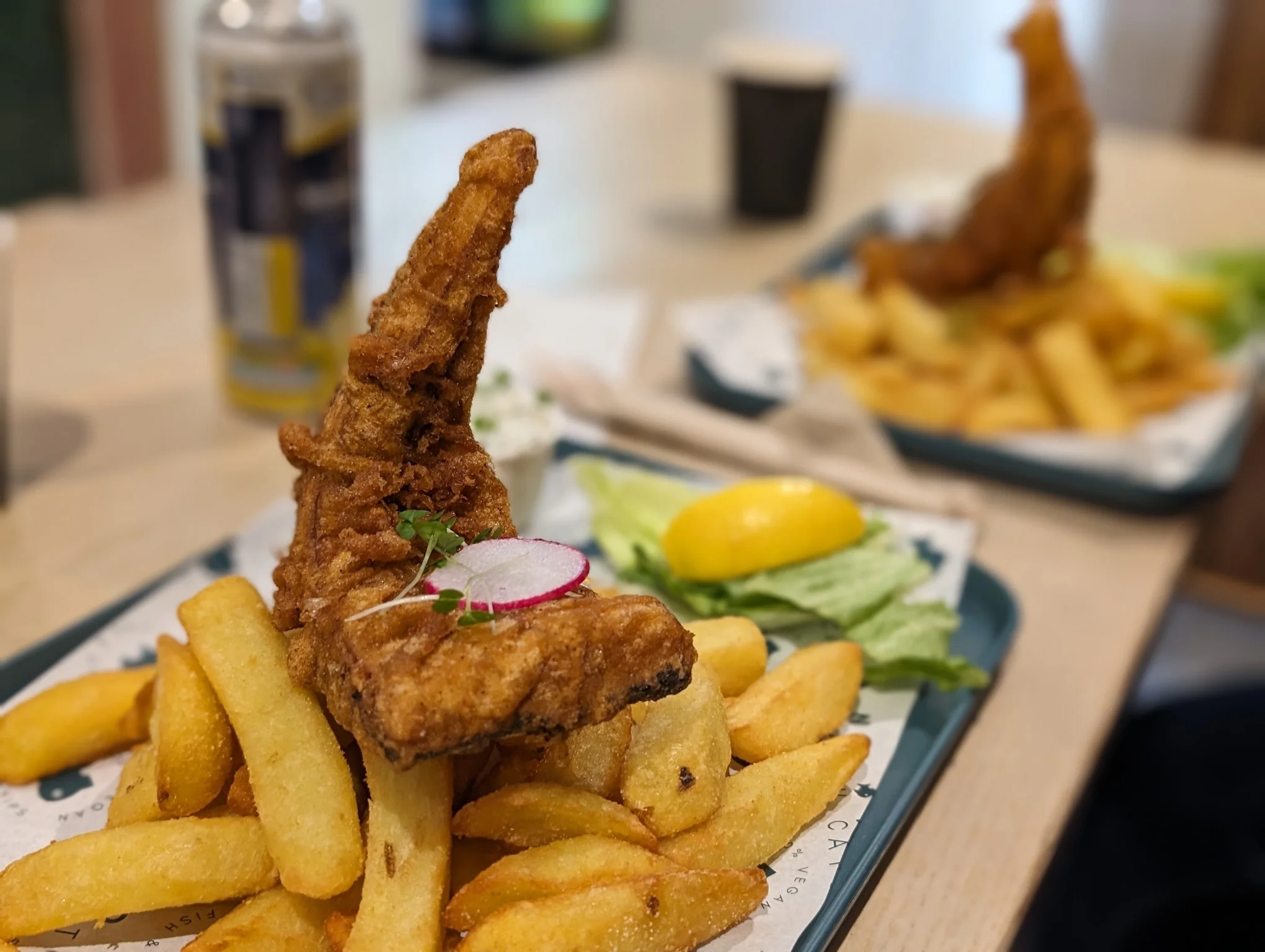 Artful photo of a large piece of battered food shaped into a curved fishtail atop a pile of chunky chips.