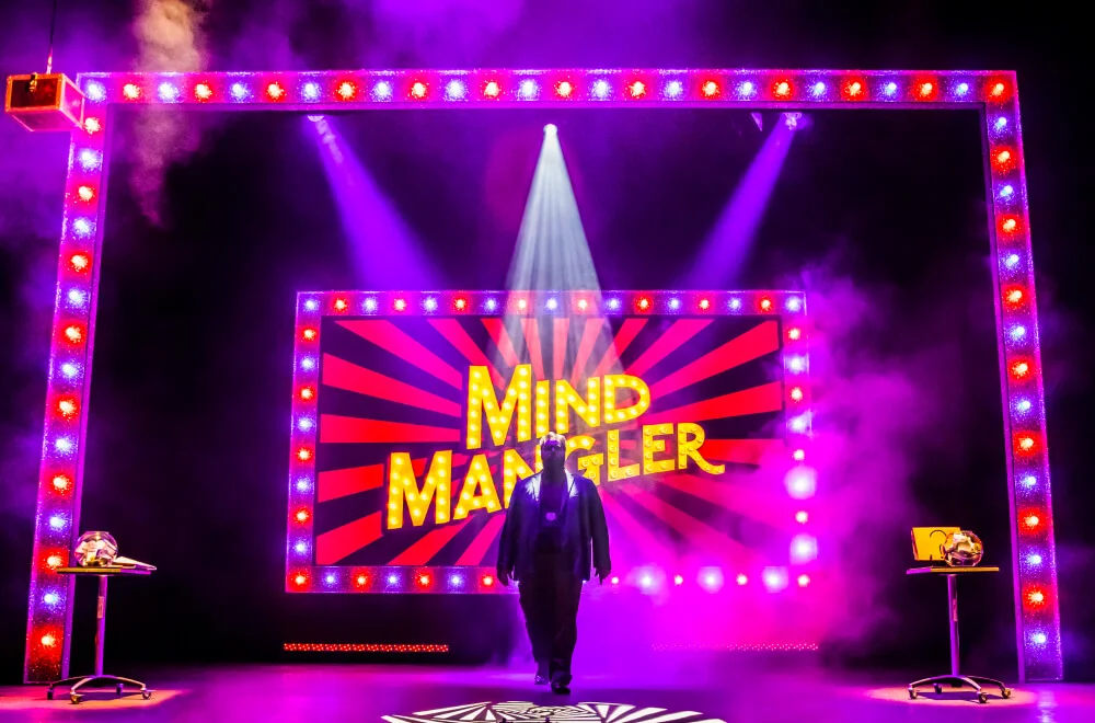 Photo of a man standing on a stage with an illuminated sign reading "Mind Mangler".