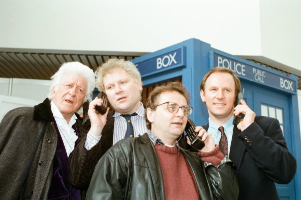 Photo of Pertwee, Colin Baker, McCoy, and Davison standing outside the TARDIS. They all have 1990s mobile phones.